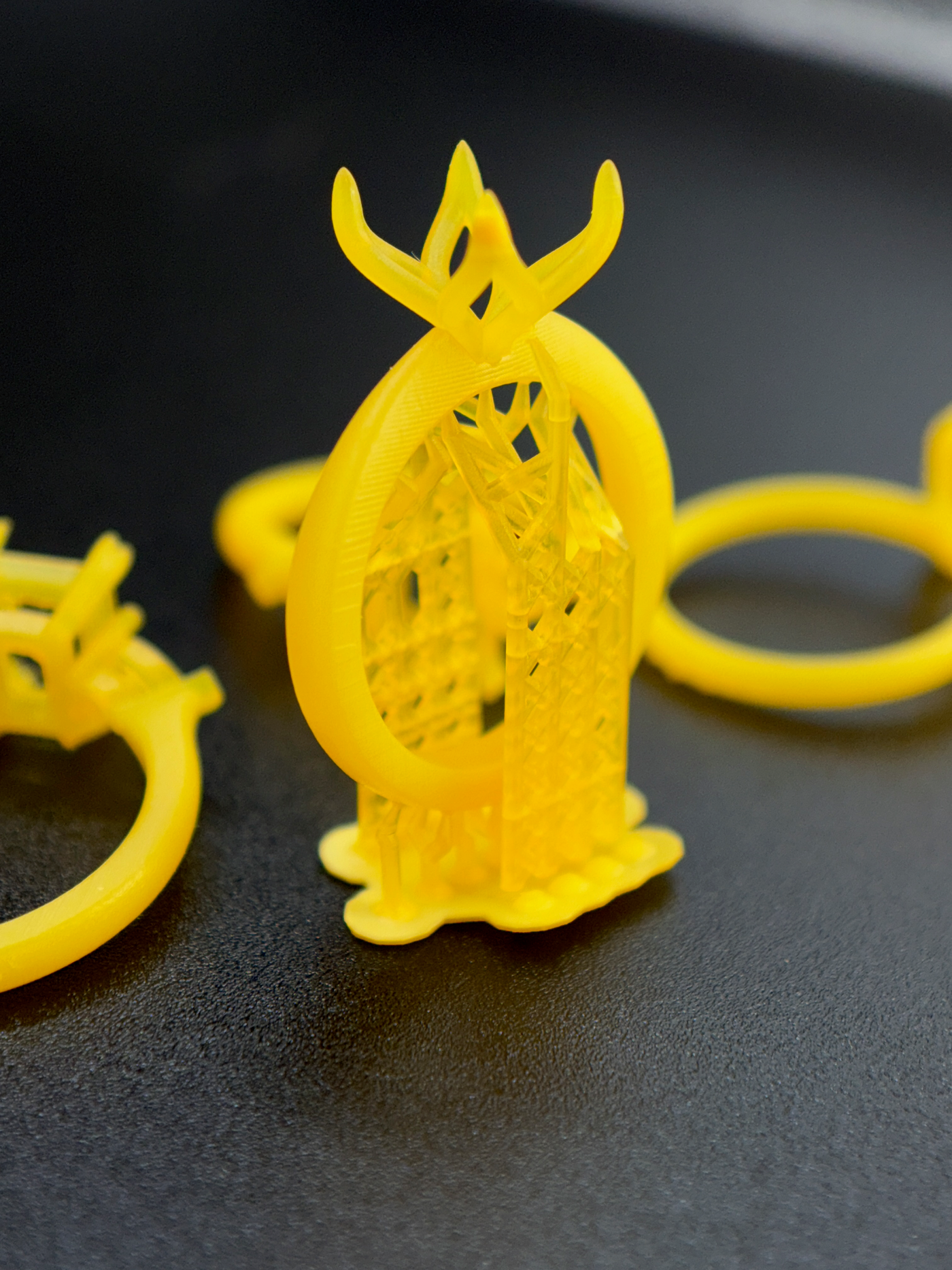A bright golden 3D-printed ring before post processing with supports, shown on a dark surface with similar golden rings blurred in the background. Printed in-house by our CTO, Erica Odom on a Phrozen Mini 8KS at 35um wit ACF film.