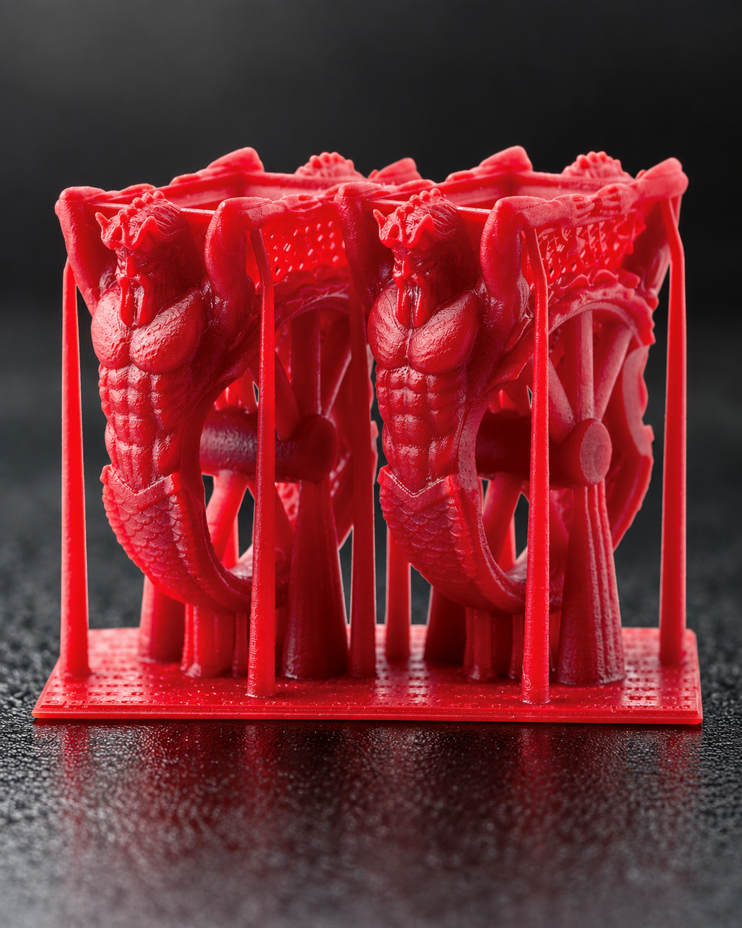 Close-up of a red 3D printed direct investment casting ring part made with SuperWAX v2 resin material. The ring features a muscular, stylized design, mounted on a matching red base plate against a dark, textured background.
